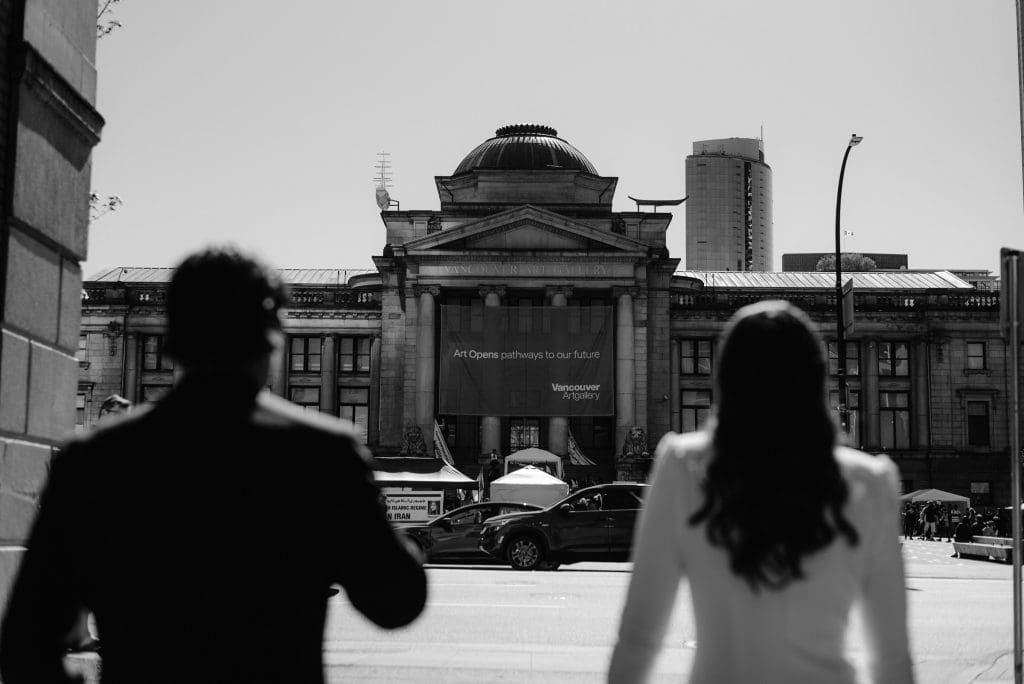 Black and white photo of bride and groom standing together, both looking at a church.