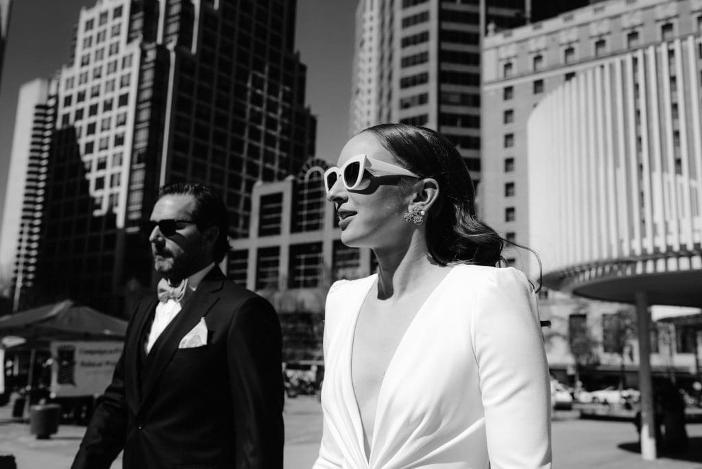 A bride in a white dress and sunglasses walks beside a groom in a black tuxedo through a city street, with tall modern buildings in the background, captured in black and white.