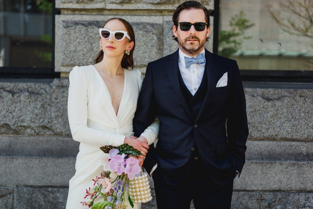 A bride in a white dress holding a bouquet stands arm-in-arm with the groom.